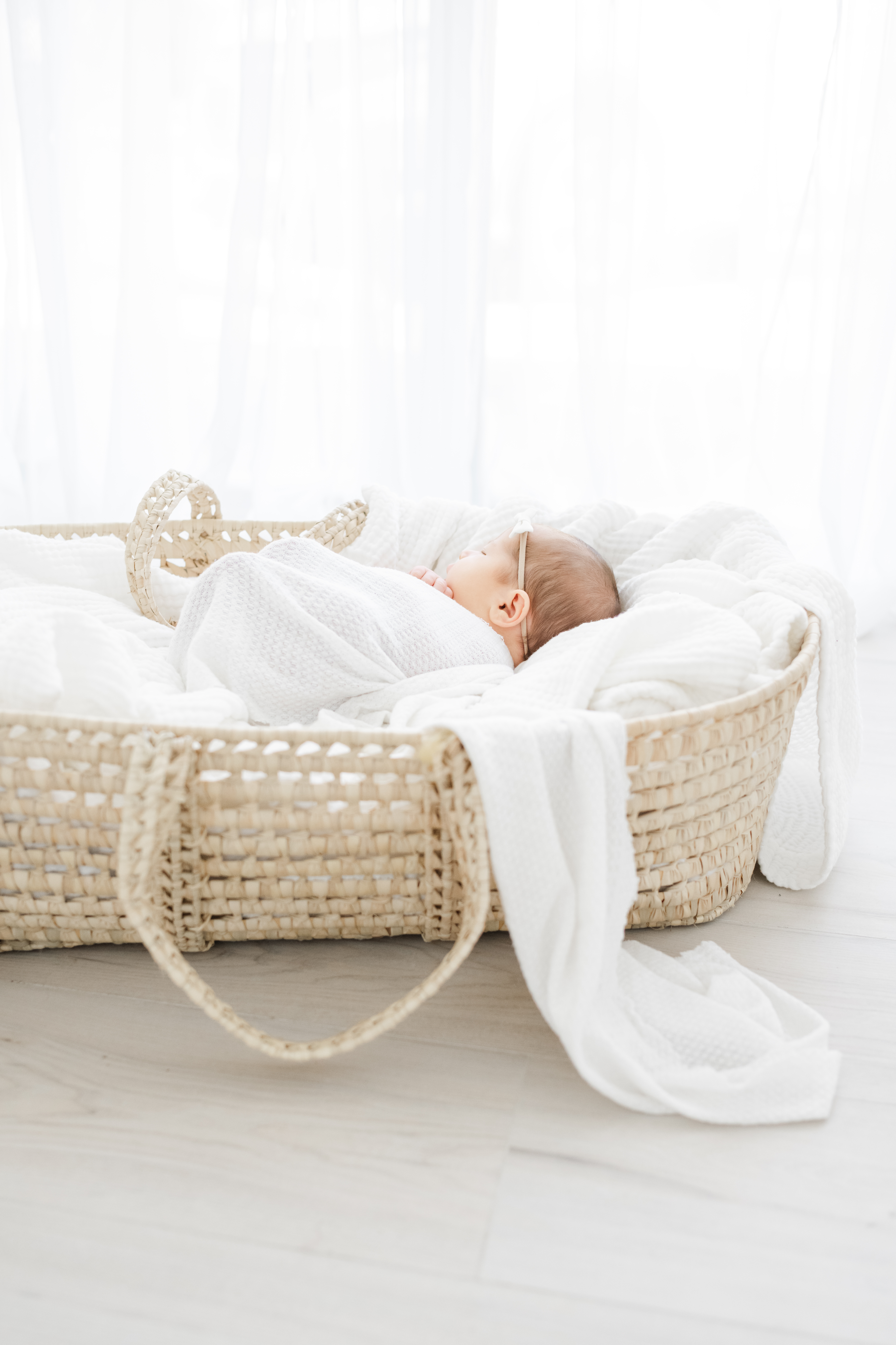 Newborn-baby-sleeping-peacefully-in-a-woven-Moses-basket-wrapped-in-a-white-blanket-with-natural-light-filtering-through-sheer-curtains-in-Rogers-Arkansas-Photography-Studio